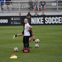 A person in sports attire stands on a soccer field, surrounded by several soccer balls and a carrier with bottles. The field is bordered by advertisements for Nike Soccer and app stores. Several people sit in bleachers in the background.